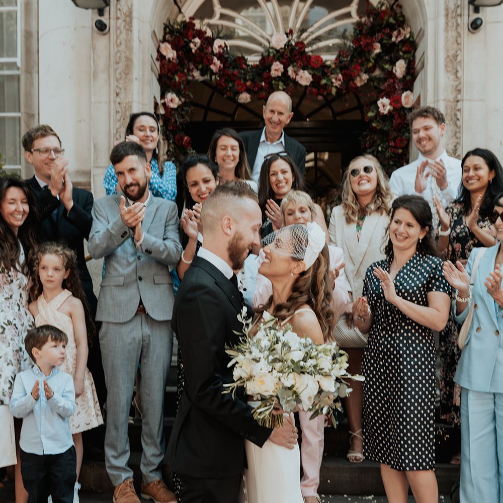 bride in a birdcage veil smiling at her groom in a black suit on the steps of Chelsea Town hall 
