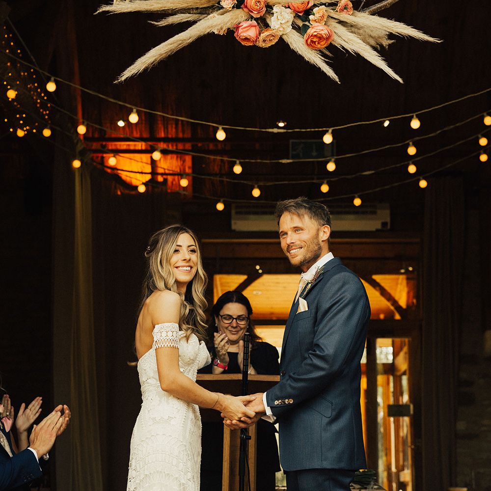 Smiling bride and groom hold hands under pampas grass and rose flower cloud 