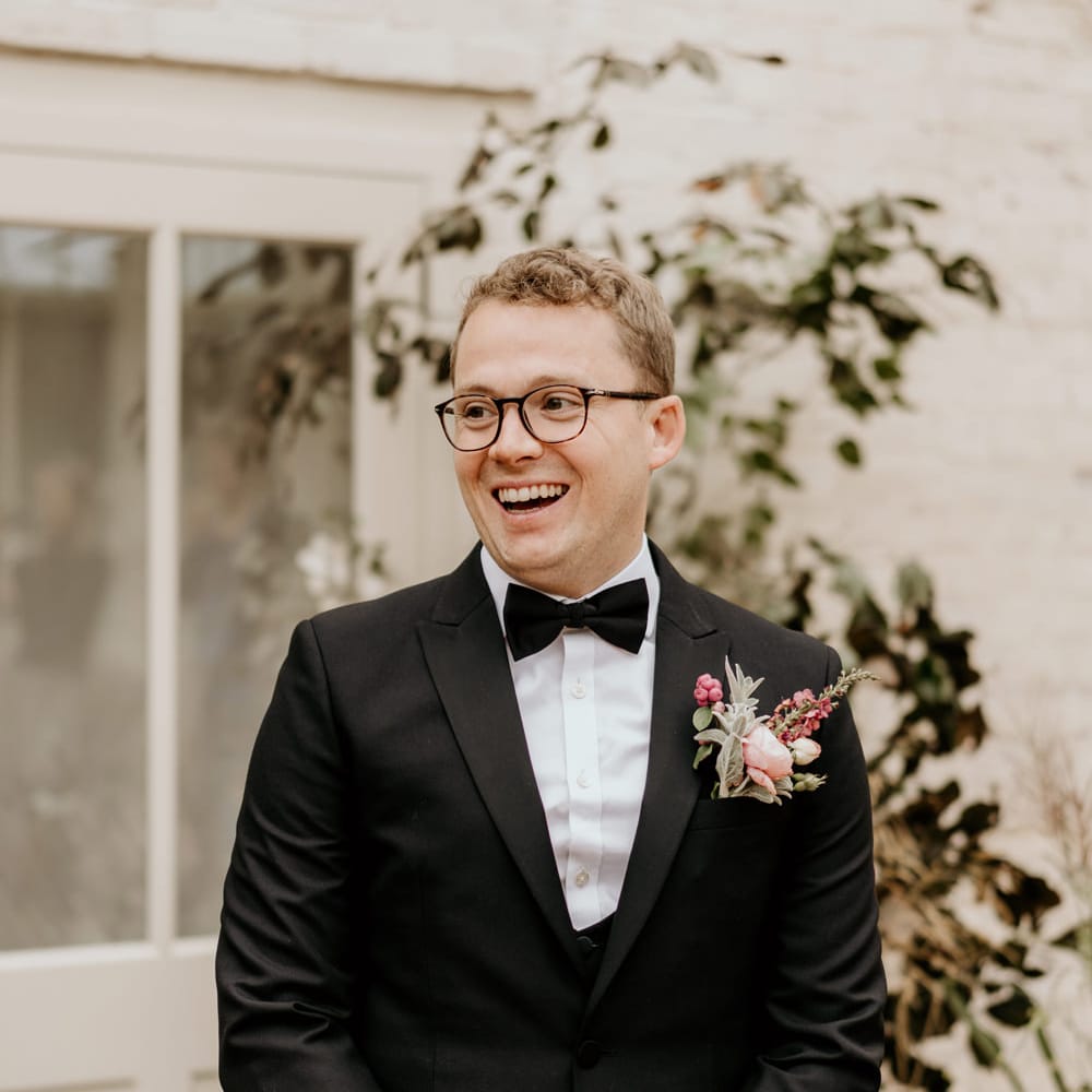 White blonde groom in glasses wearing a black tuxedo and bow tie with a pink flower buttonhole 