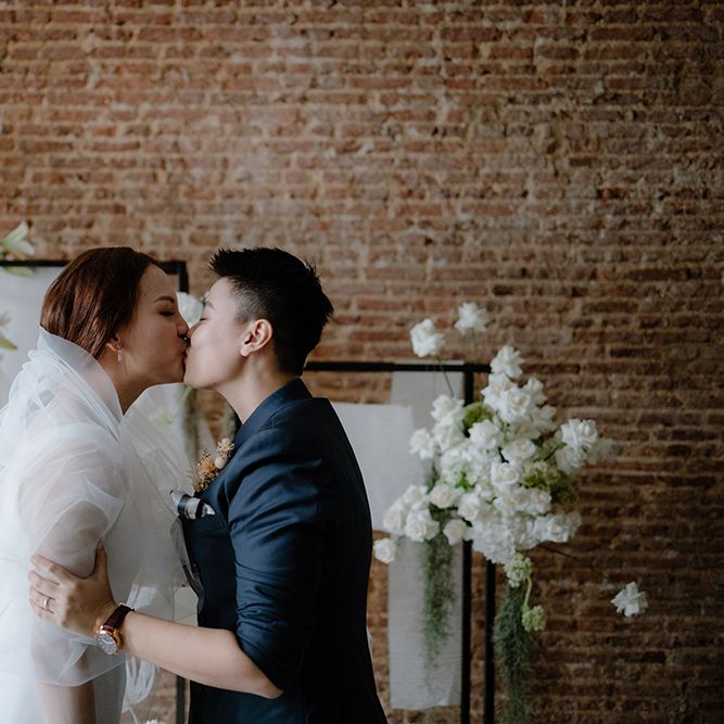 Asian Lesbian wedding ceremony with bride in a navy suit kissing her bride in a white minimalist dress and veil