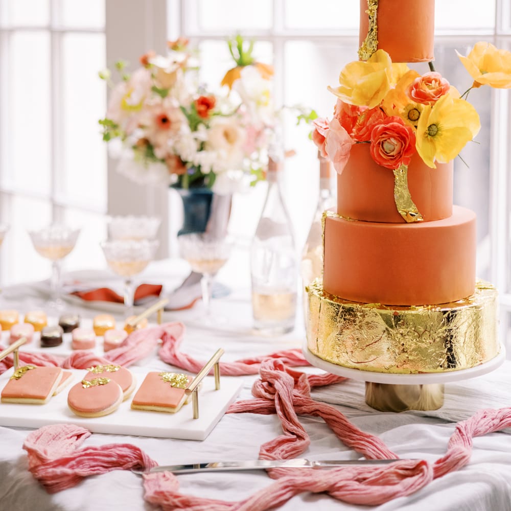 Four tier wedding cake with varying colours of orange frosting, complete with floral decor and gold leaf