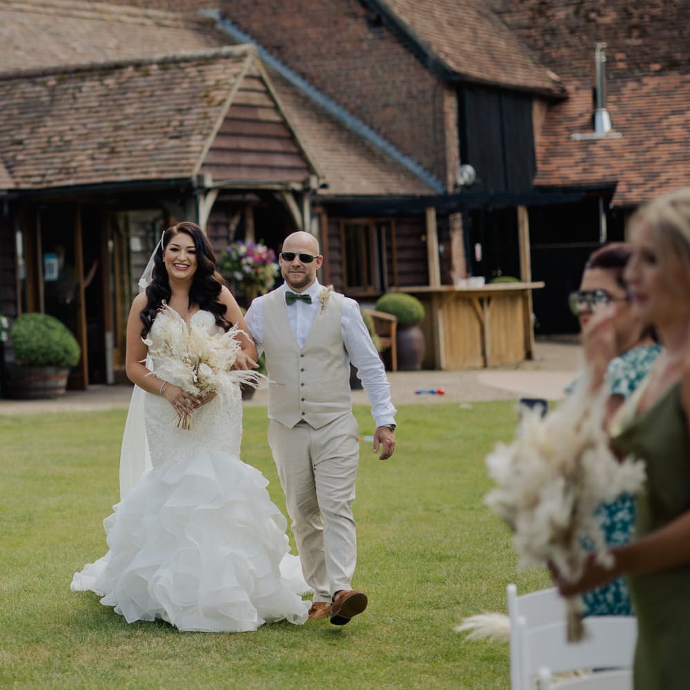 Bride in a fishtail strapless wedding dress walking down the aisle at outdoor wedding ceremony at Cooling Castle Barn