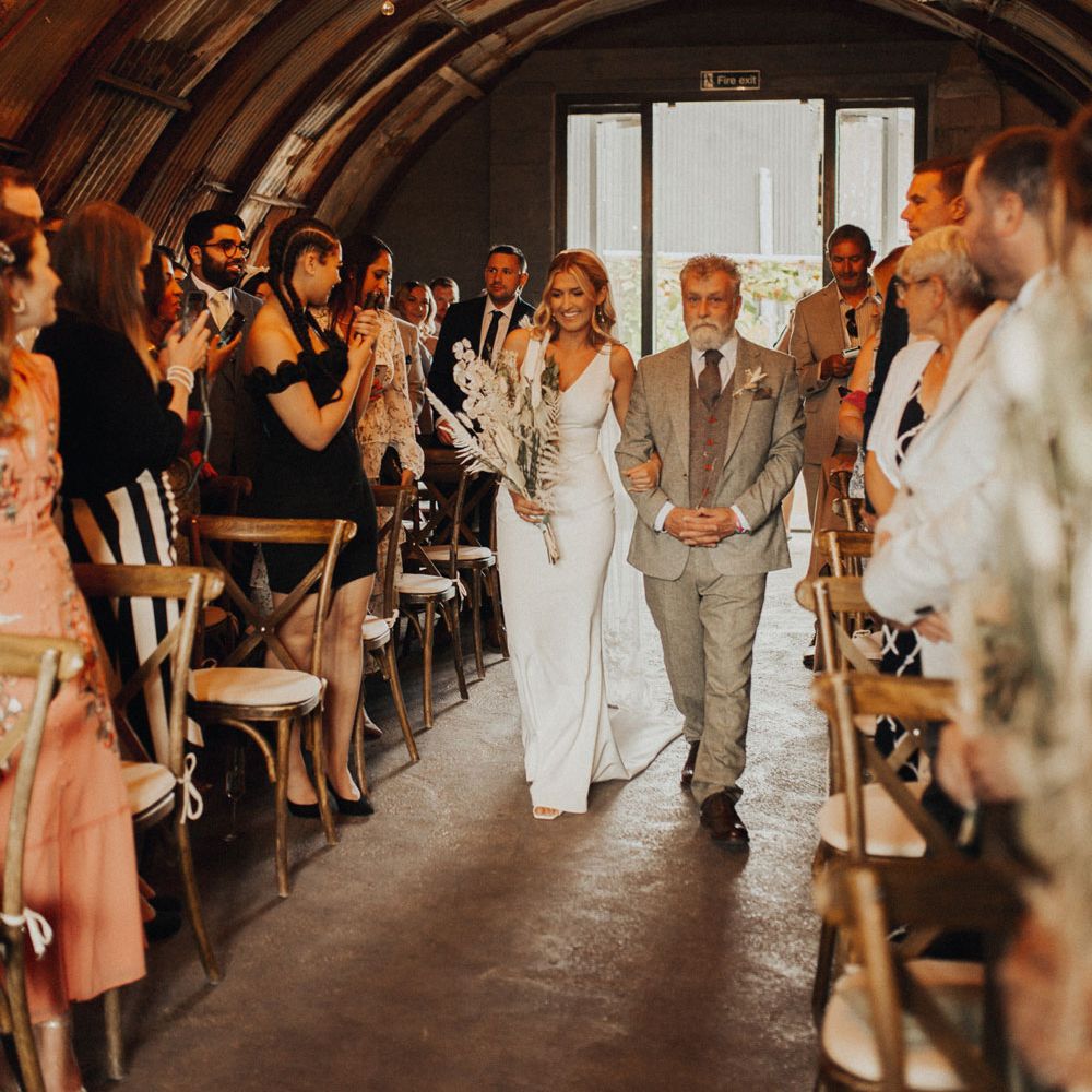 Smiling bride in white V neck Made With Love wedding dress and veil walks up the aisle arm in arm with man in sage green suit in wedding ceremony room lit up with chandeliers as guests watch at Anran Devon