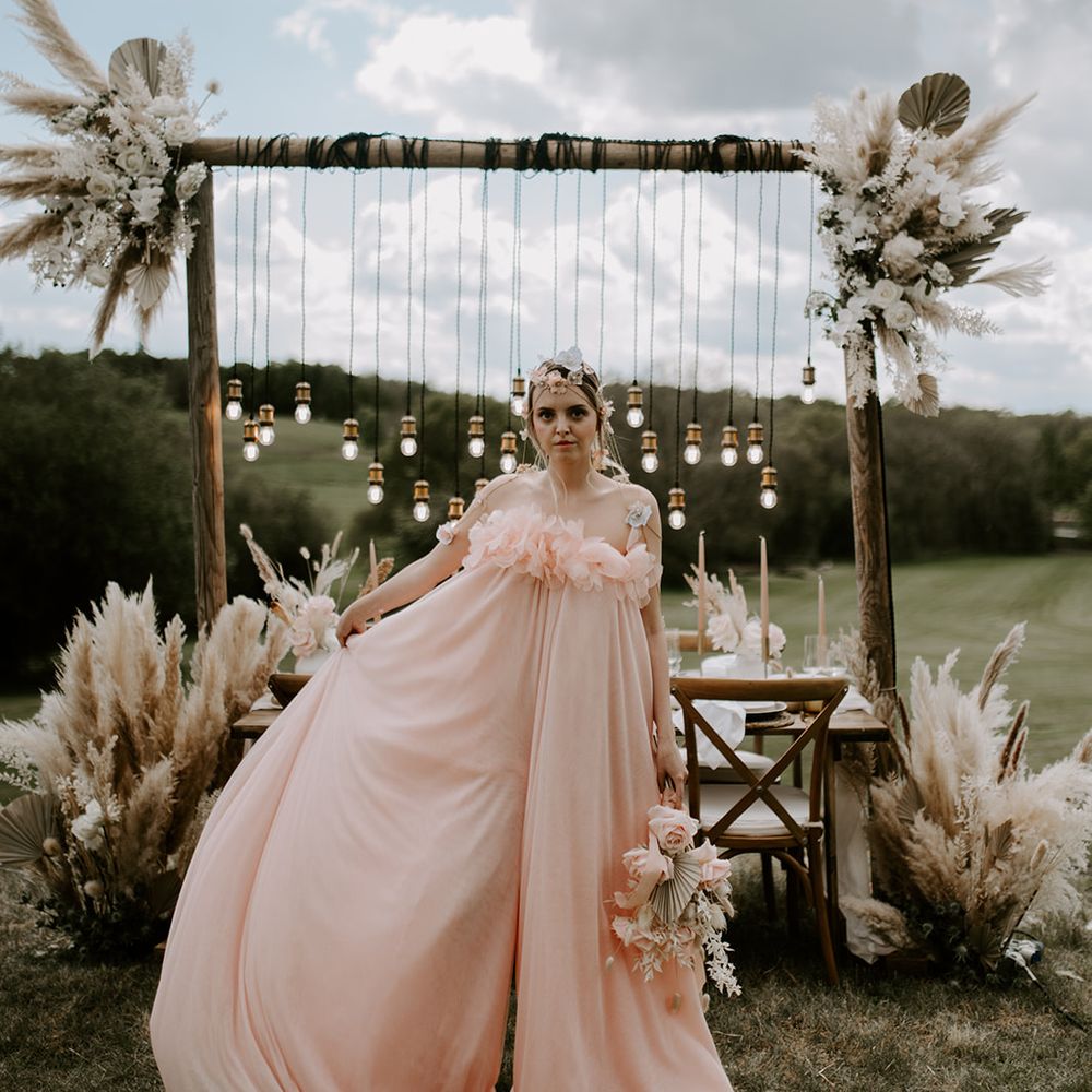 Boho bride in a blush pink jumpsuit with appliqué flowers wearing a chain headdress and standing in front of an outdoor reception table with light installation and dried flower decor 