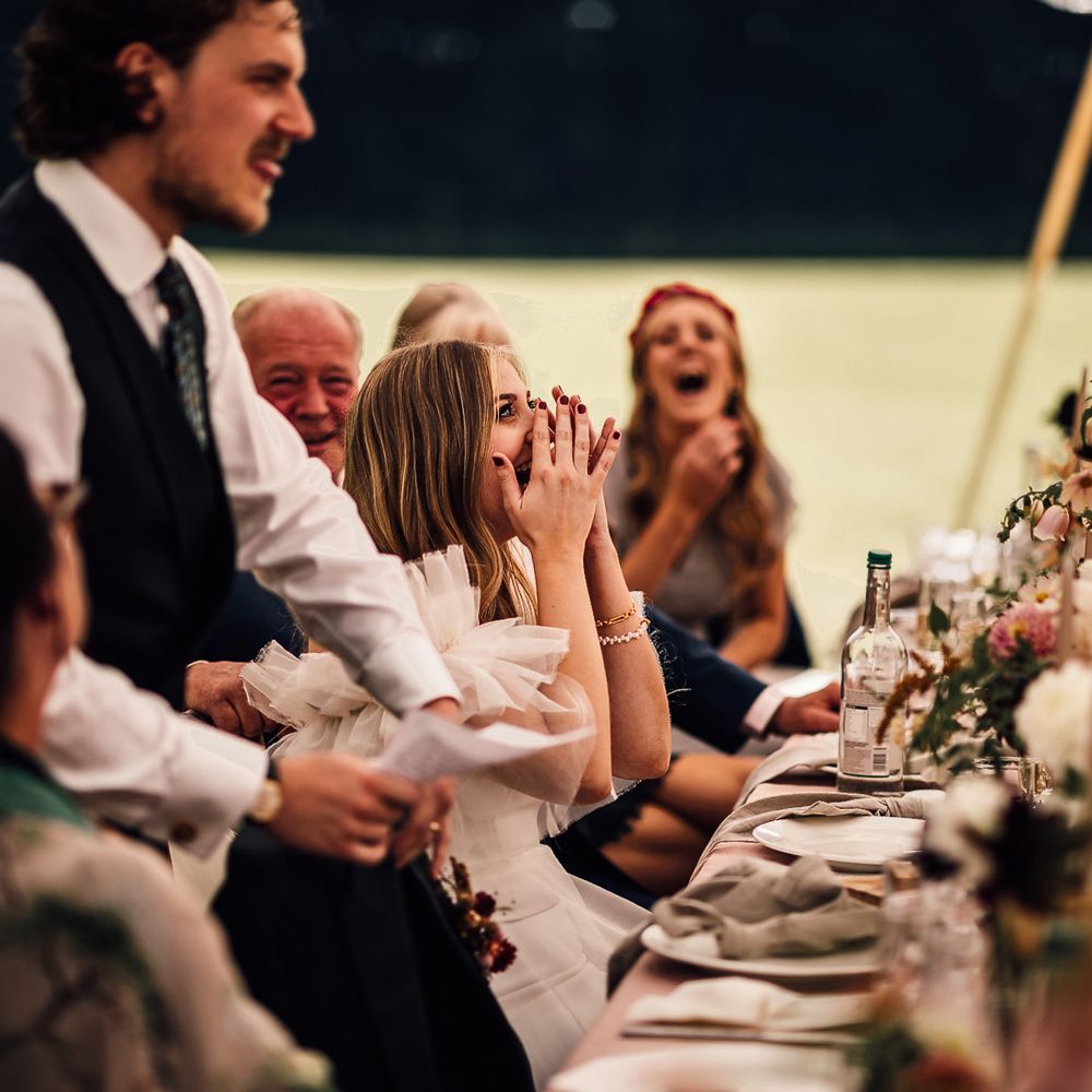 Bride in white shirt, blue waistcoat and patterned tie makes speech during garden marquee wedding reception in Cornwall as bride in Halfpenny London Mayfair dress and wedding guests laugh