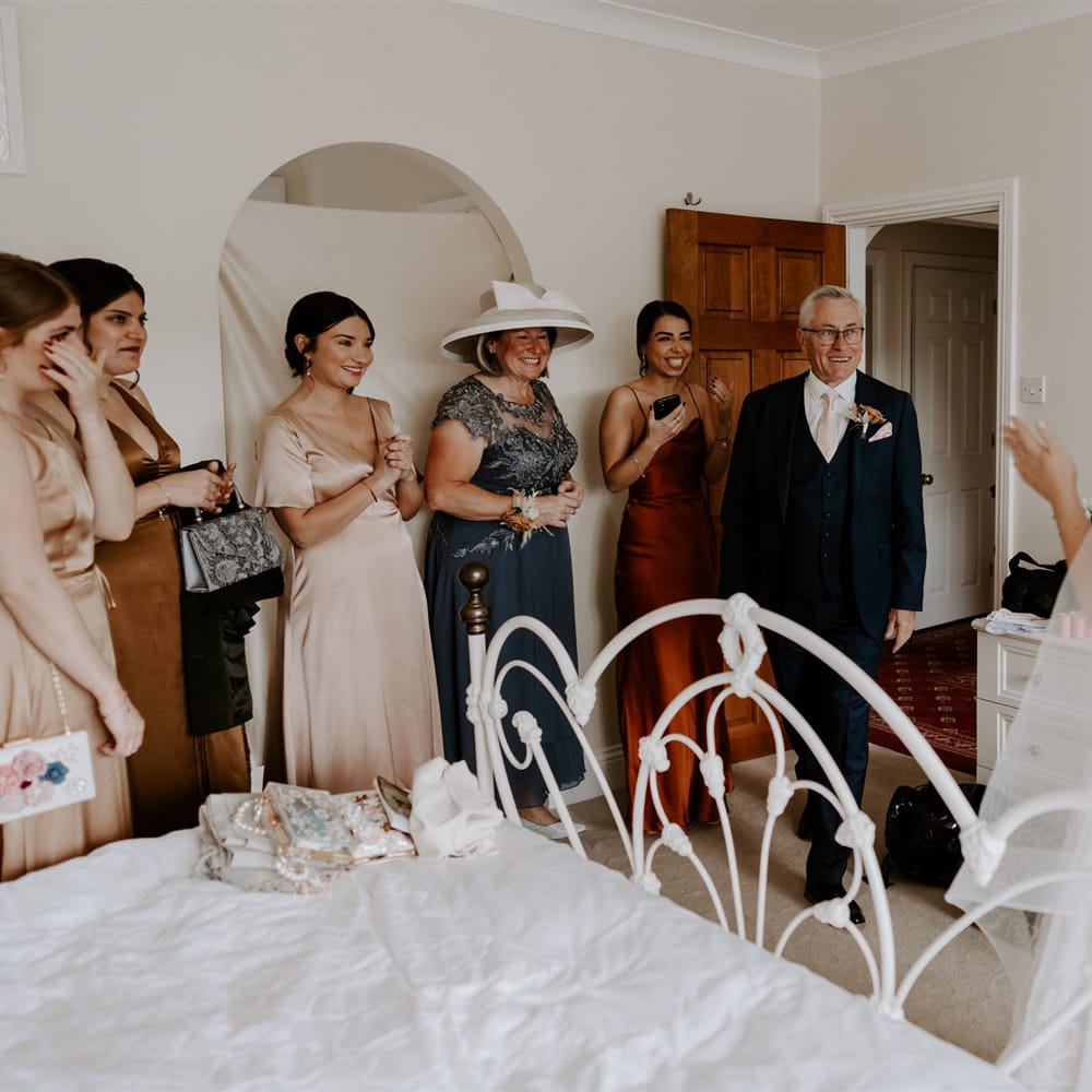 Wedding guests stand in bedroom and smile at bride in homemade lace wedding dress and veil before DIY garden wedding ceremony in Bedfordshire
