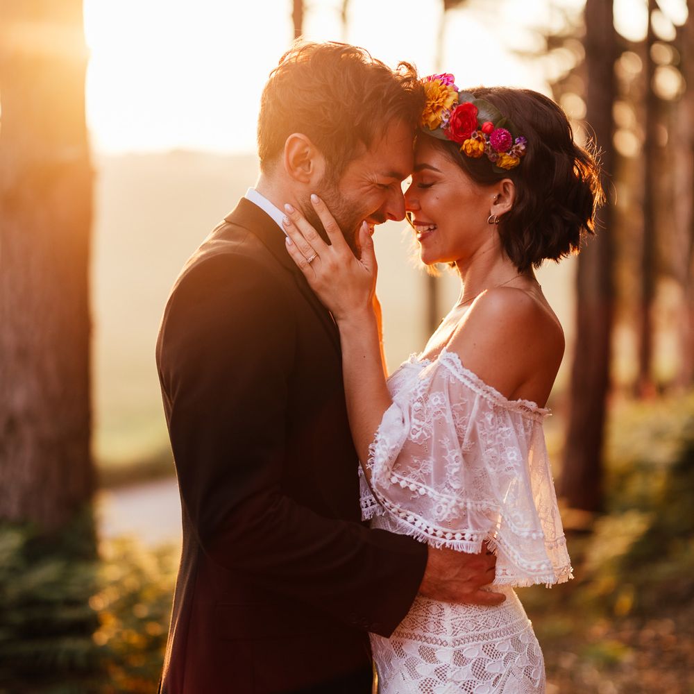 Smiling bride in off the shoulder Grace Loves Lace wedding Dres sand flower crown stands with groom in black suit in woodland during golden hour