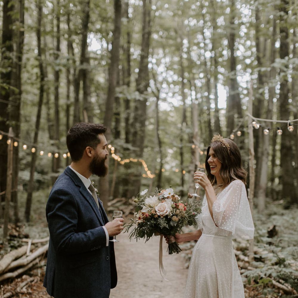 Bride & groom laugh and drink champagne with one another in woodlands at The Dreys wedding venue