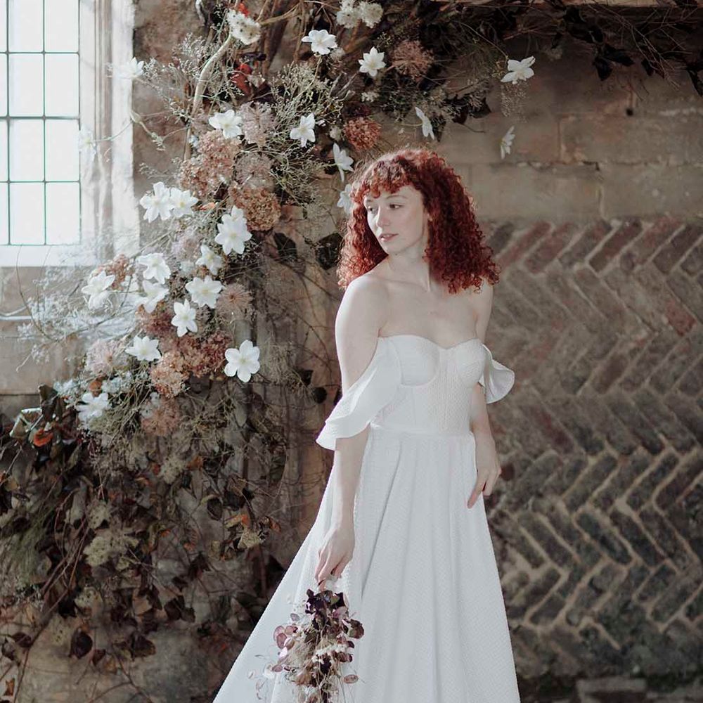 Bride red curly hair in a strapless princess wedding dress holding a minimalist wedding bouquet by a dried flower floral arrangement 