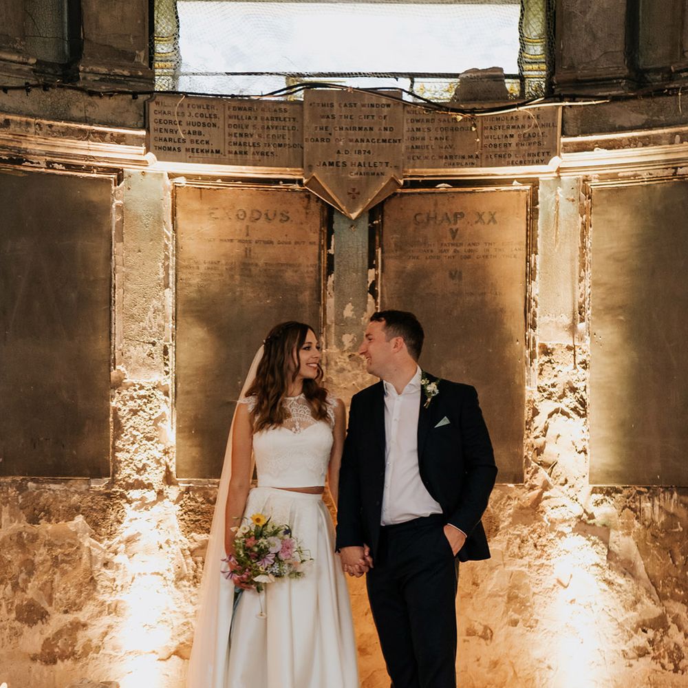 Bride in two piece wedding set and groom in navy suit stand together at their industrial style wedding venue in London 