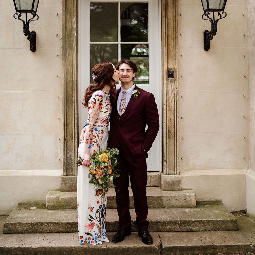 Bride in colourful wedding dress kisses the groom on the cheek wearing burgundy suit 
