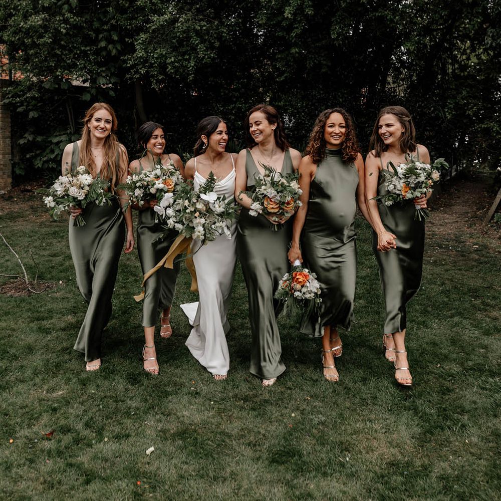 Bridal party in sage green satin bridesmaid dresses holding white and orange flower bouquets with green foliage 