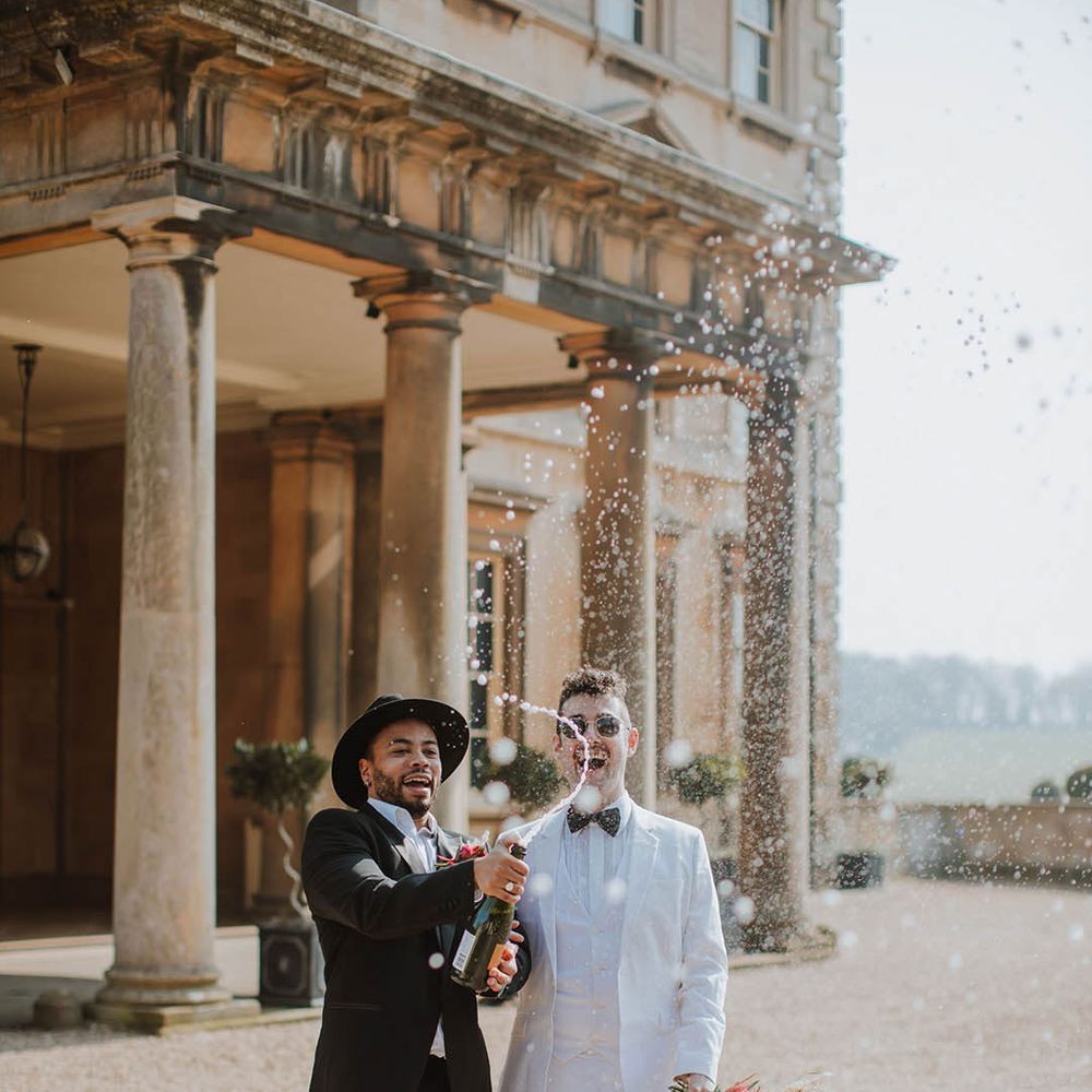 Grooms in white tux and black tux doing a champagne pop