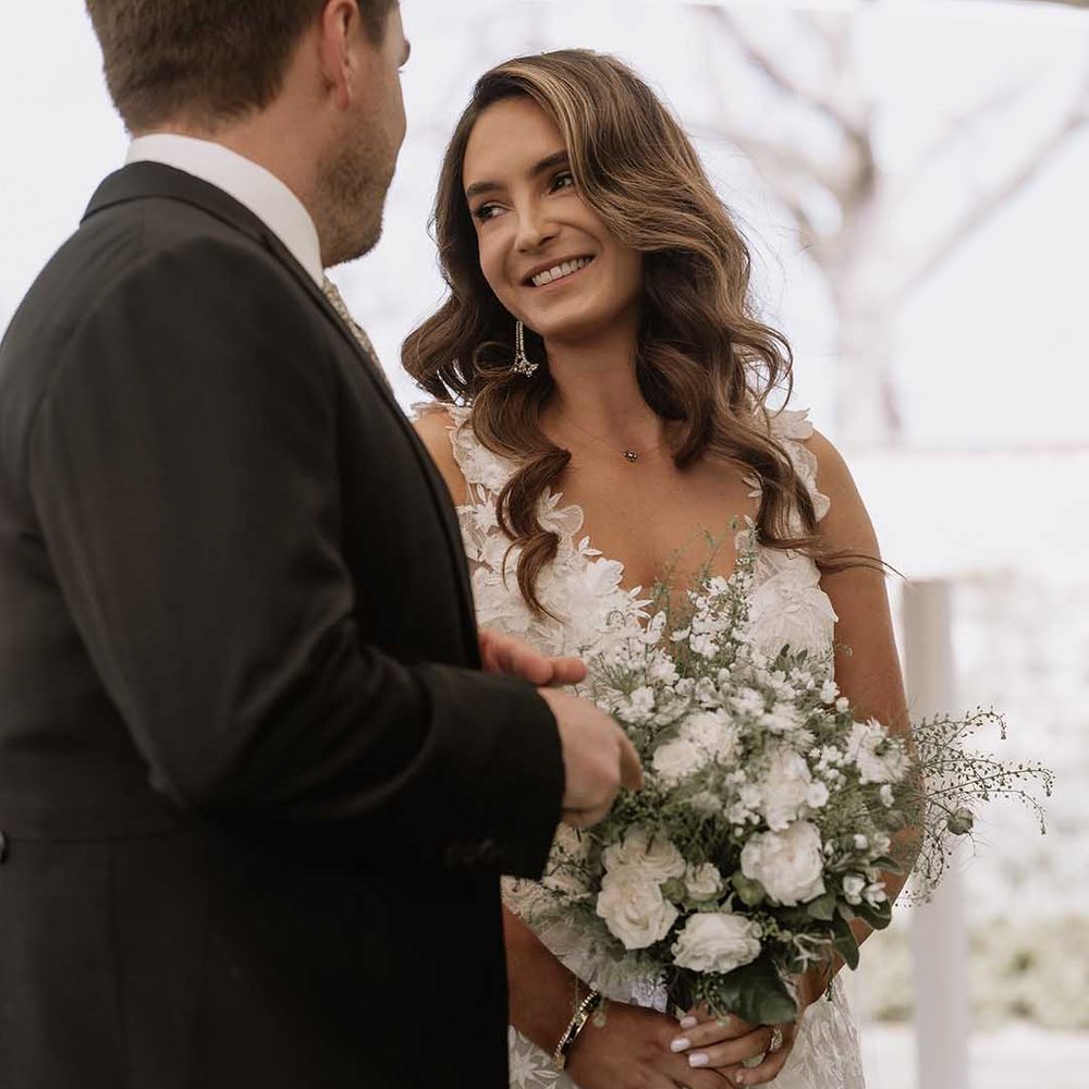 Bride holds white flower wedding bouquet with nude wedding nails and gold bracelet for classic marquee wedding as she stands at the altar with the groom 