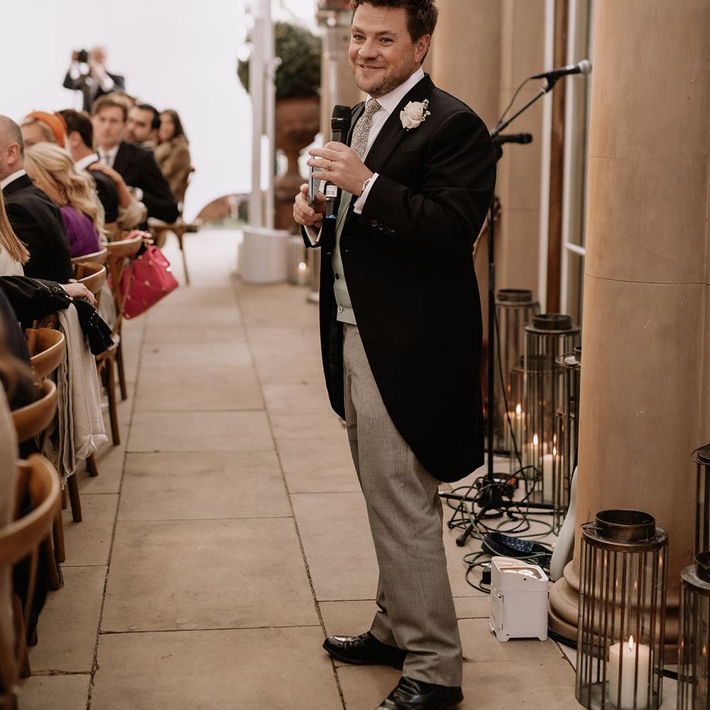 Grooms stands with microphone as he reads out his speech in morning suit 