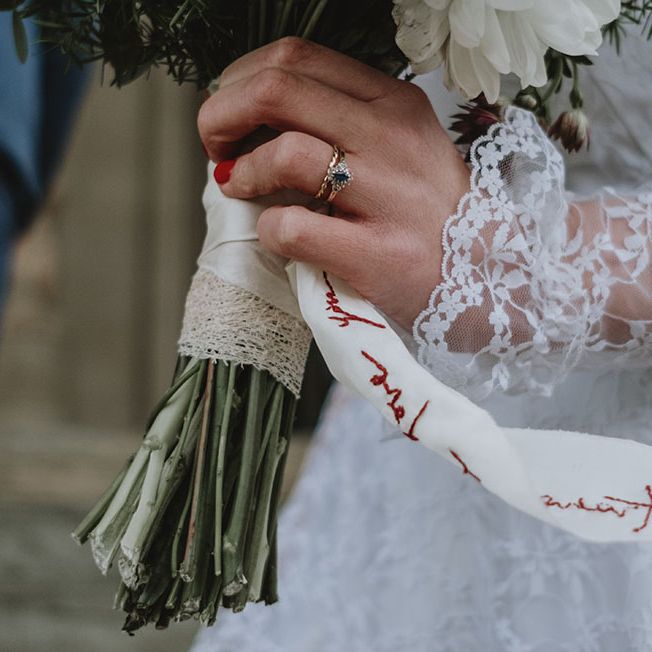 Bride wears long sleeve lace wedding dress and vintage blue diamond engagement ring whilst carrying floral bouquet tied with personalised ribbon