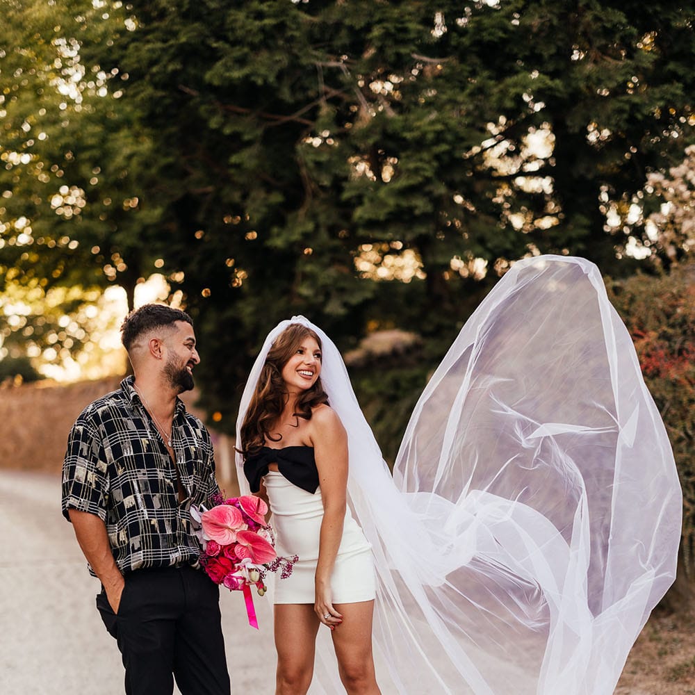 Bride wears white mini dress with black bow and throws out her veil beside her as her groom watches on 