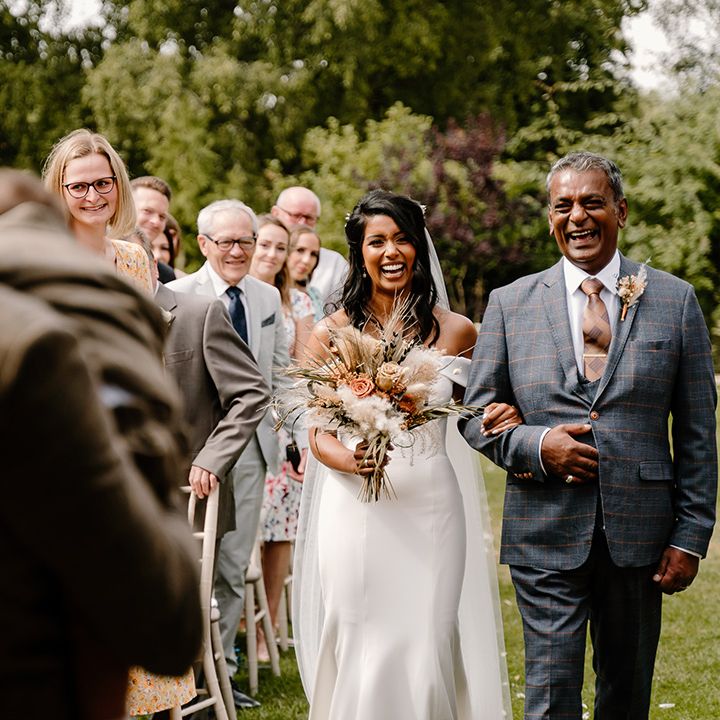 Father of the bride in a blue suit and brown tie walks the bride down the aisle in an off the shoulder Madi Lane wedding dress