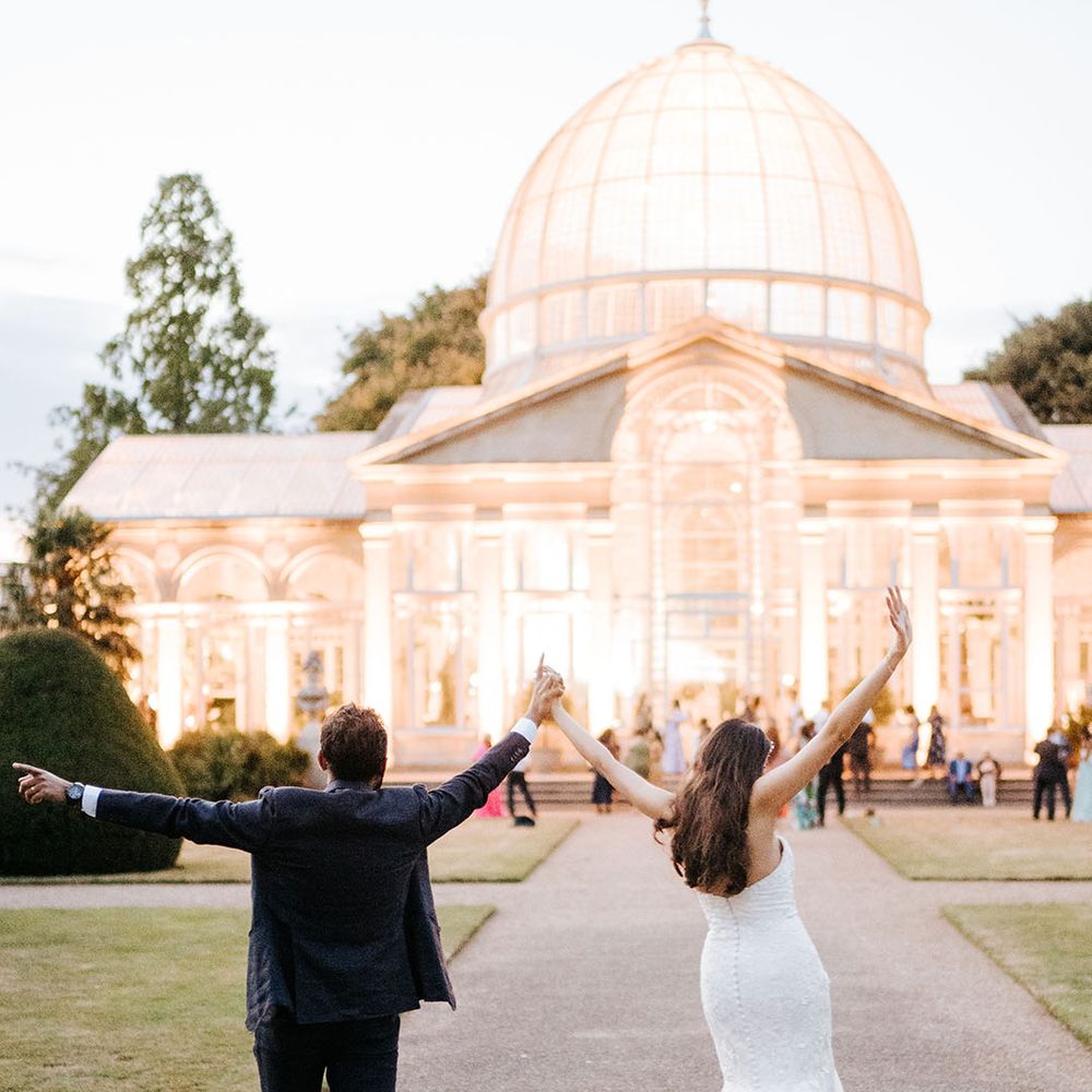 Bride & groom lift their arms in the air in celebration as they walk toward the brightly lit Great Conservatory 