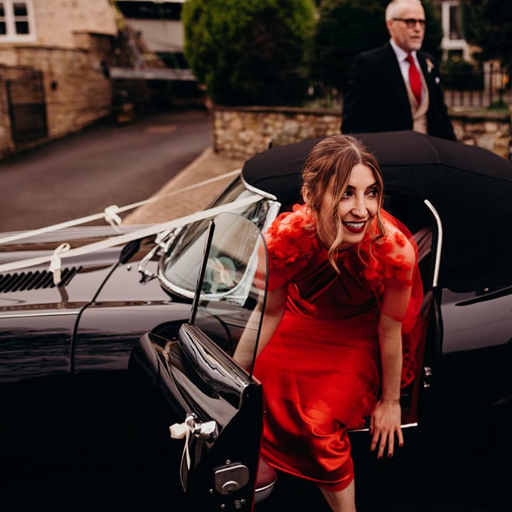 Bride in red satin wedding dress with cape with 3d appliqué roses getting out of classic convertible wedding car