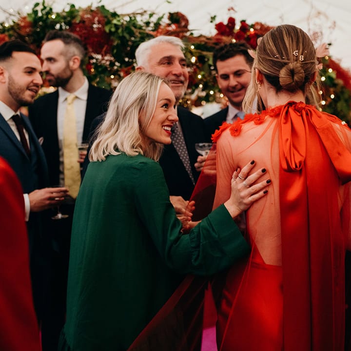 Bride in red silk wedding dress with cape, large red embellished head band, letter hair clips and large silk bow and wedding guests chatting 
