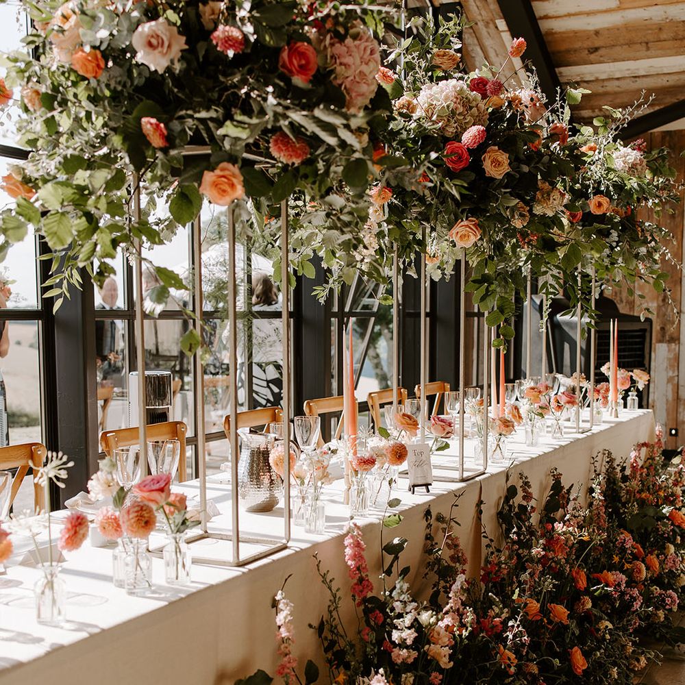 Brightly coloured floral installations line banquet tables with white tablecloth for barn wedding reception 