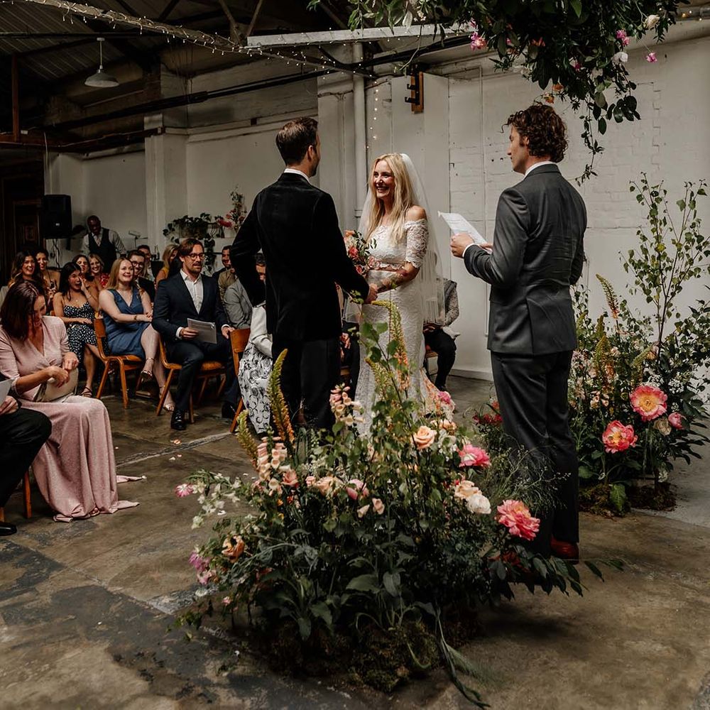 Bride and groom smiling at each other at the alter with large foliage arch decorations with colourful floral details and fairy lights
