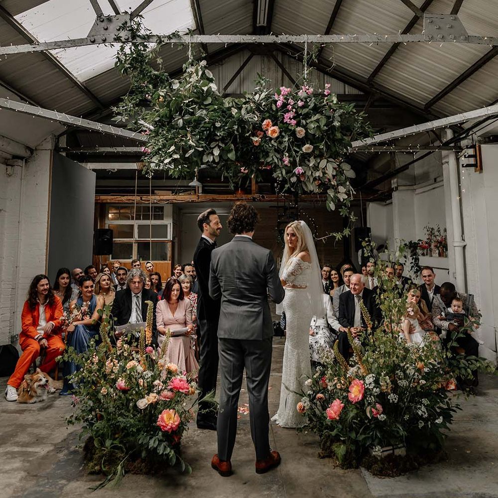 Bride and groom embracing at alter with large foliage arch decorations with colourful floral details and fairy lights