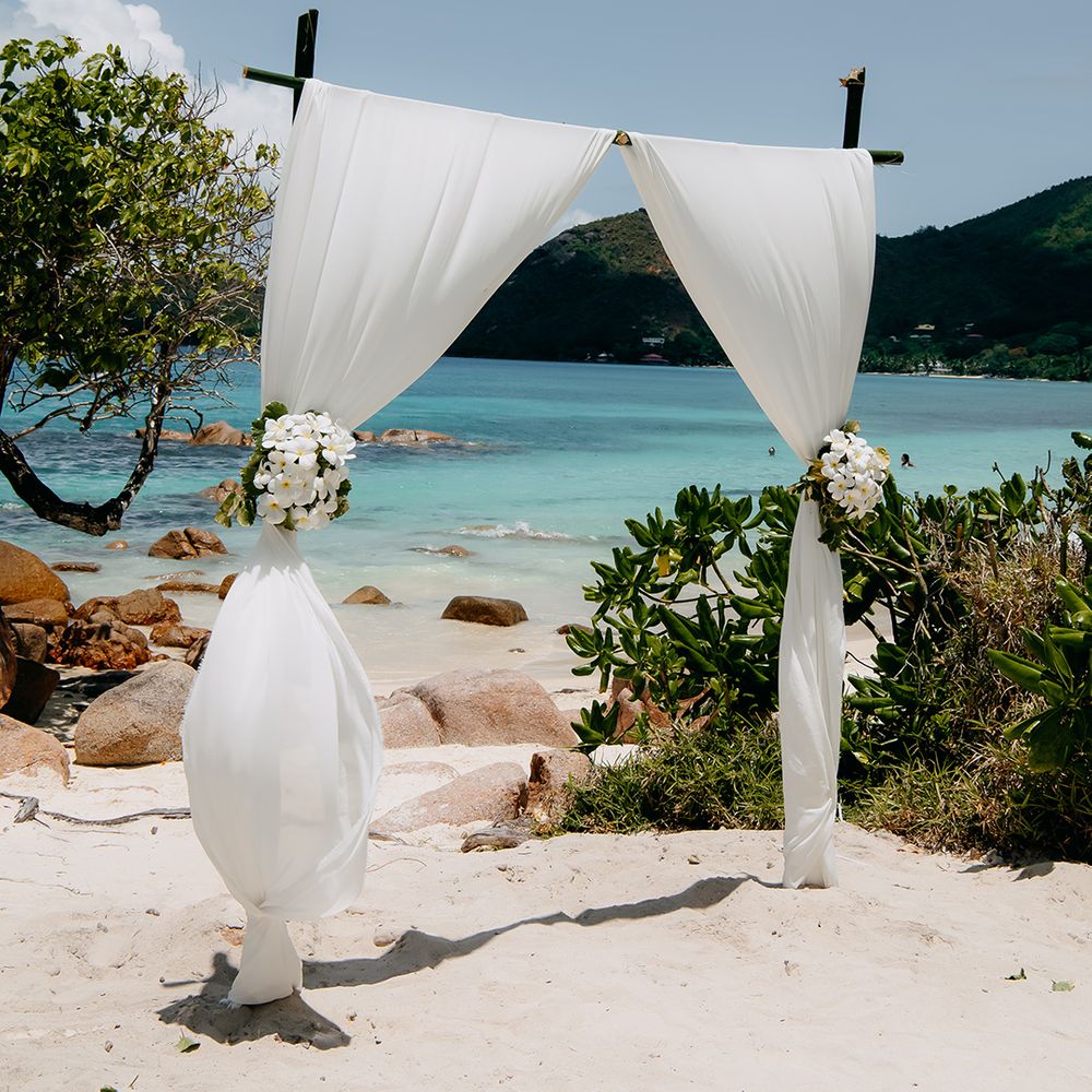 White chiffon fabric wedding arch drapes with white tropical flower arrangements on the beach in Seychelles