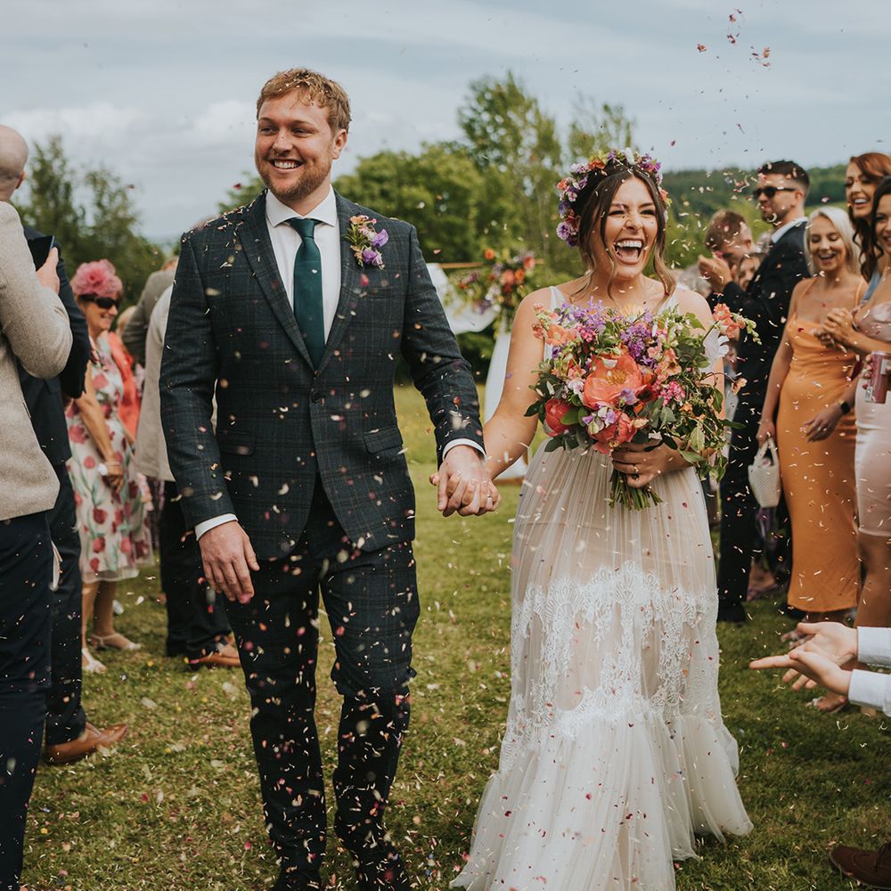 Bride carrying colourful bouquet walks through colourful confetti exit with her groom