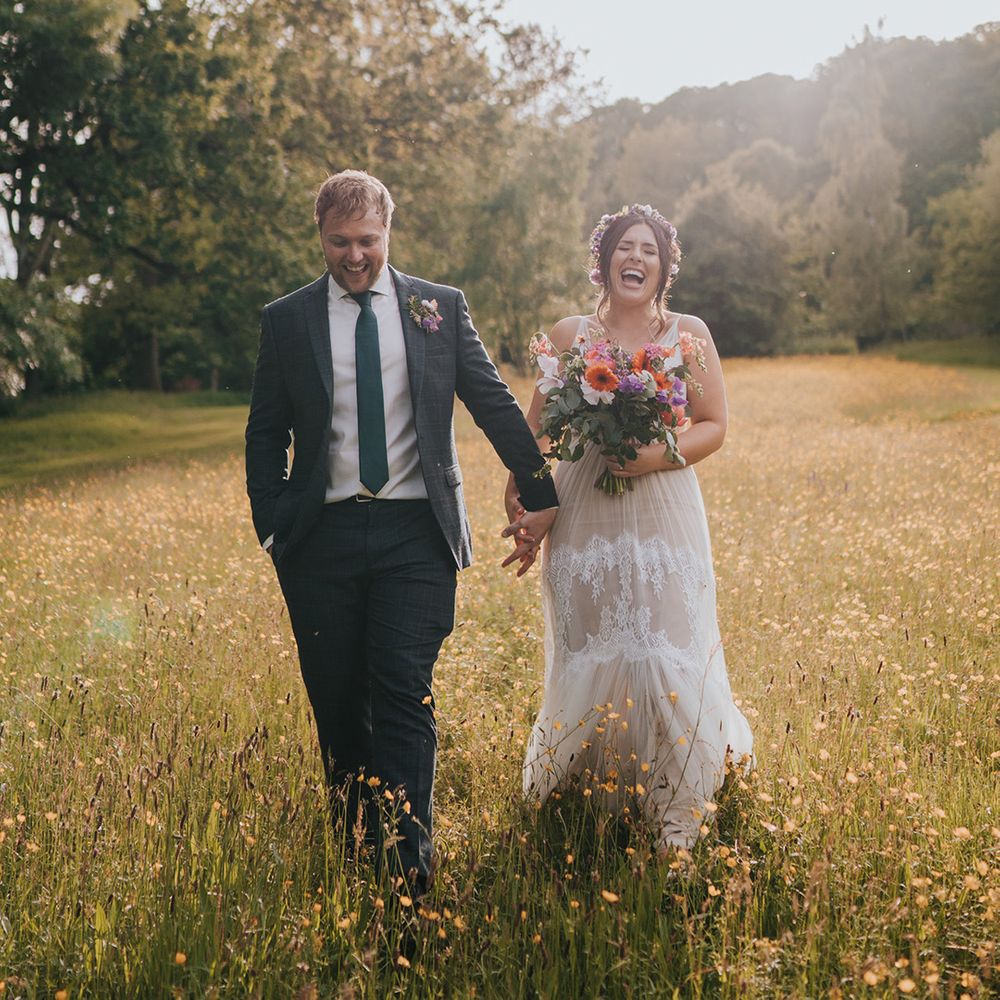 Bride holding colourful bouquet walks through fields with her groom during golden hour couples portraits 
