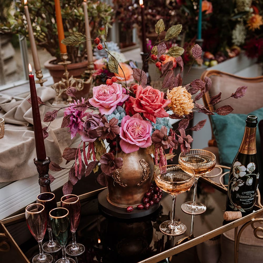 Black glass and gold drinks cart with champagne glasses and cocktail glasses, a bottle of champagne, and a single tier iced wedding cake in the style of an aged golden vase illed with handmade wafer paper florals