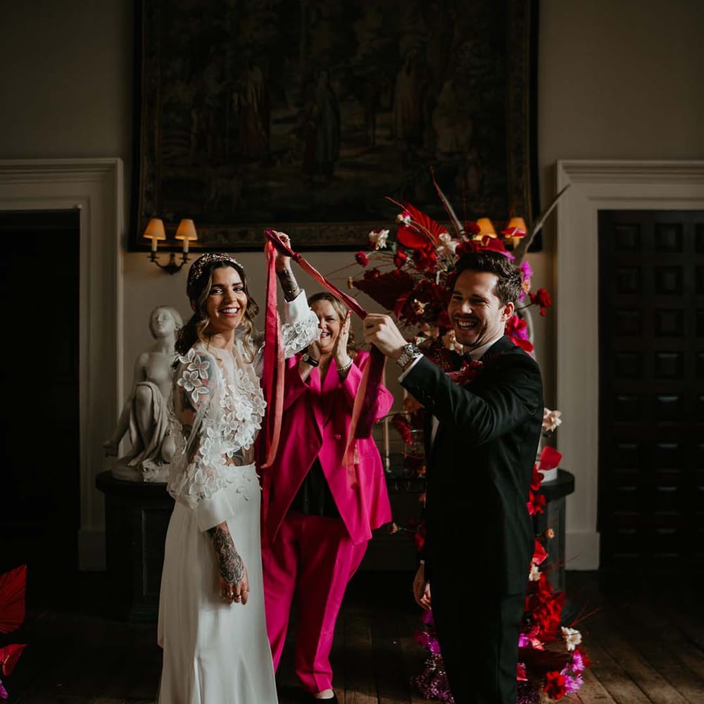 Groom in classic black tuxedo with magenta amaryllis and orchid boutonniere and bride in two piece bridal separates with puff sleeve sheer crop top with 3d appliqué flowers holding magenta handfasting ribbon at Elmore Court 