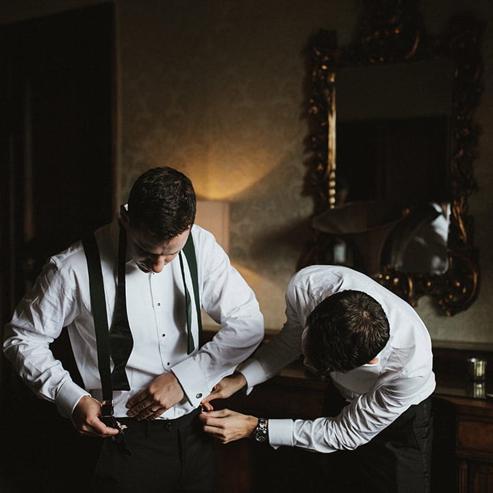Groom and groomsman getting ready together in white shirts, braces, and bow ties with black trousers for a traditional wedding 