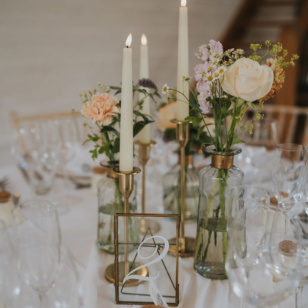 Clear glass table number sign on gold photo frame with white taper candles in gold candle holders and pastel wedding flowers in bud vases 