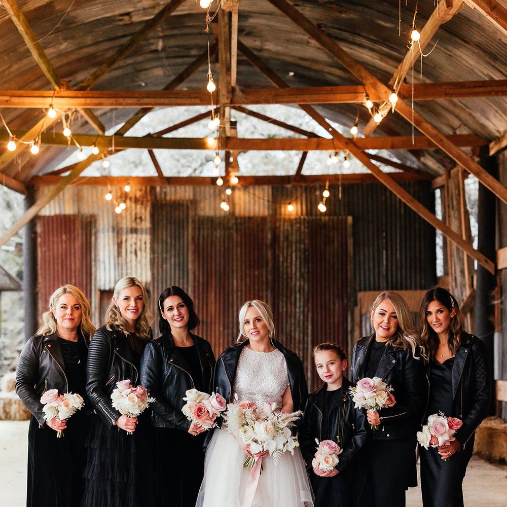 Bridesmaids with side swept hair in black bridesmaid dresses and black leather jackets with the bride in bridal separates with pale pink rose bouquets