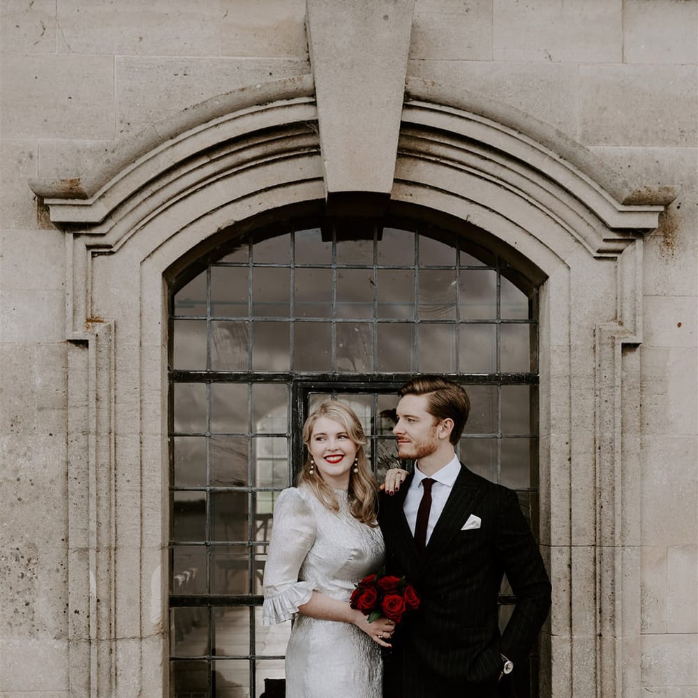 The bride wearing red lipstick, pearl earrings and silver wedding dress holding red rose bouquet with the groom in a pinstriped suit 