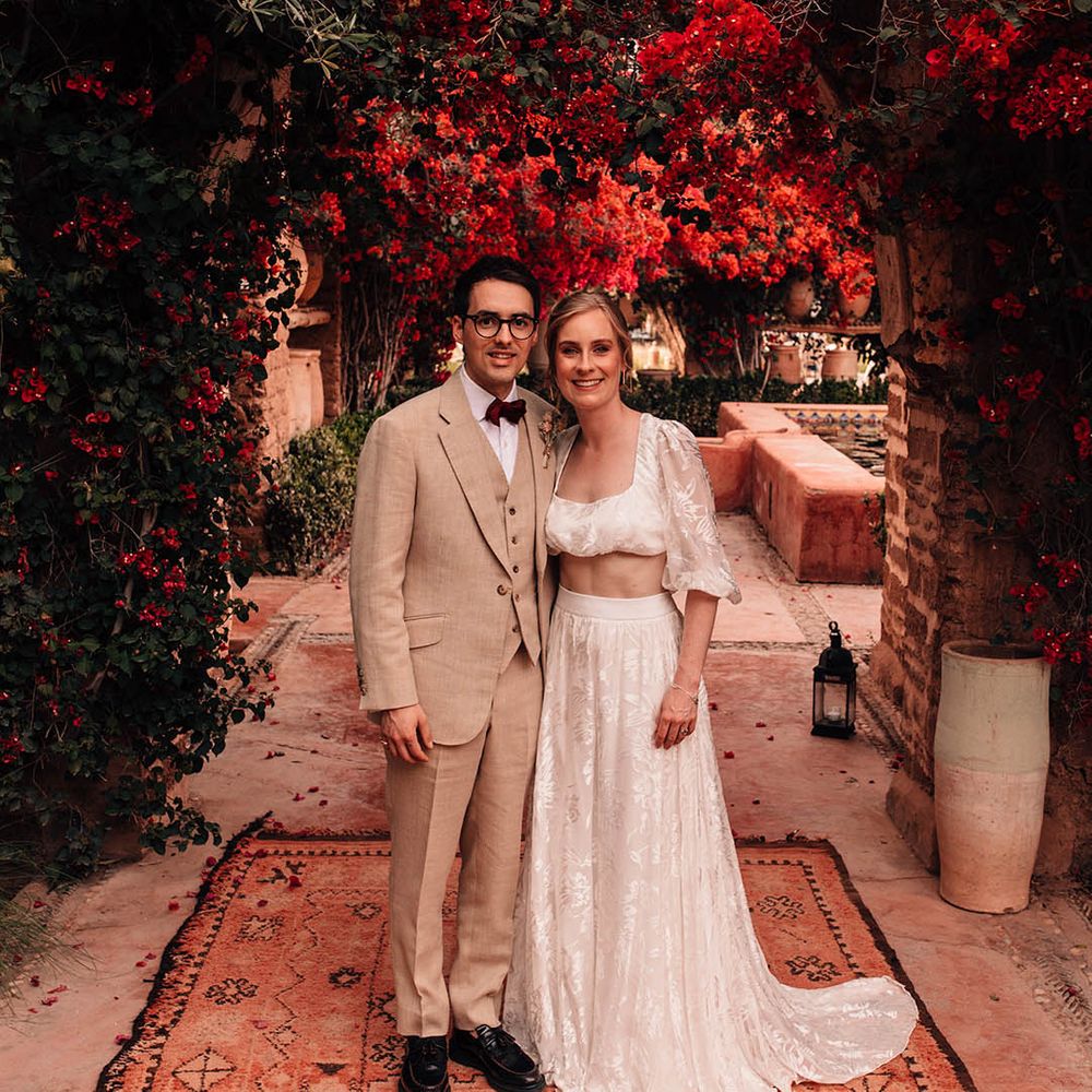 Groom in cream three piece suit with red bow tie and dried flower buttonhole with the bride in modern two piece set 