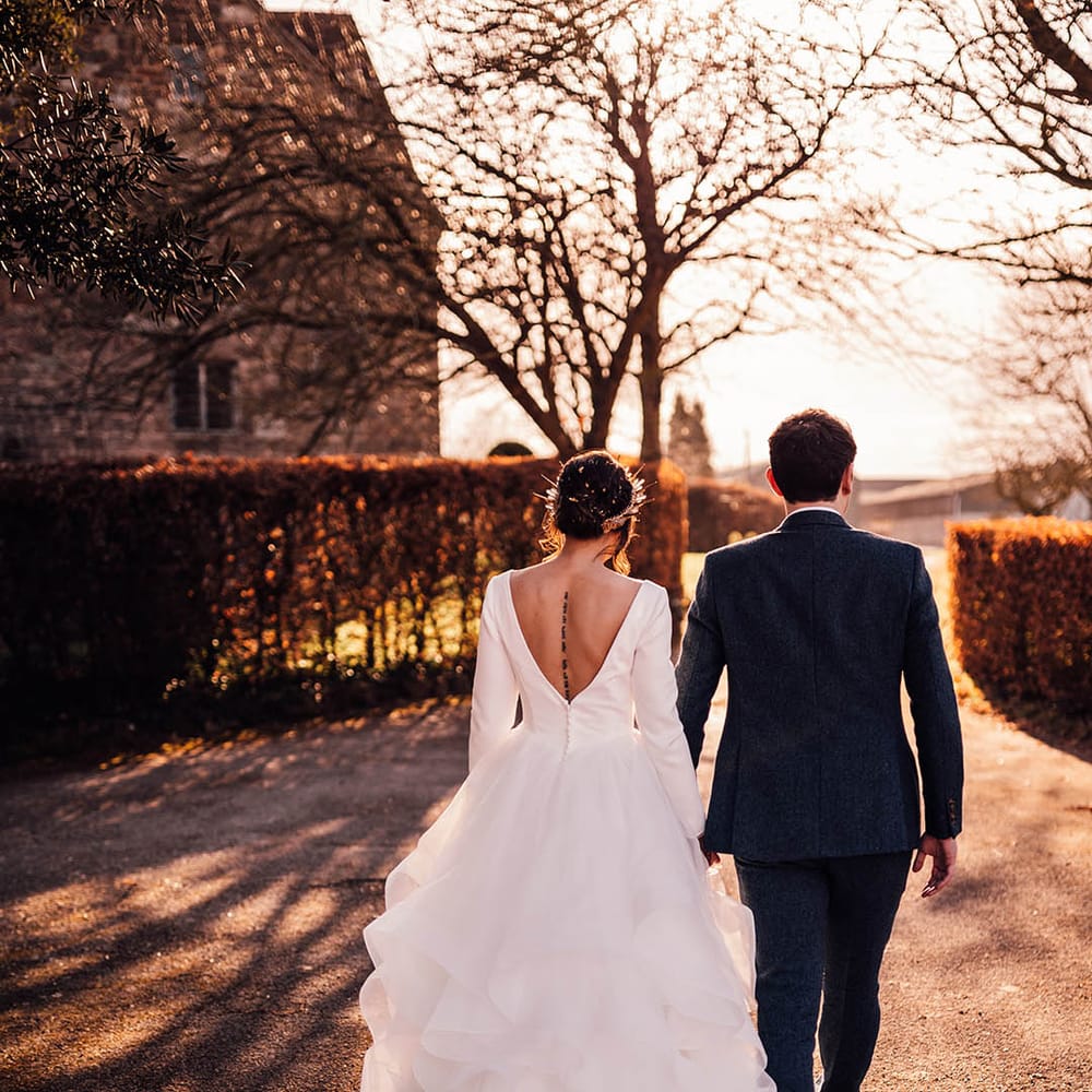 Bride in low back dress with full princess skirt walking around their wedding venue with groom in navy suit 