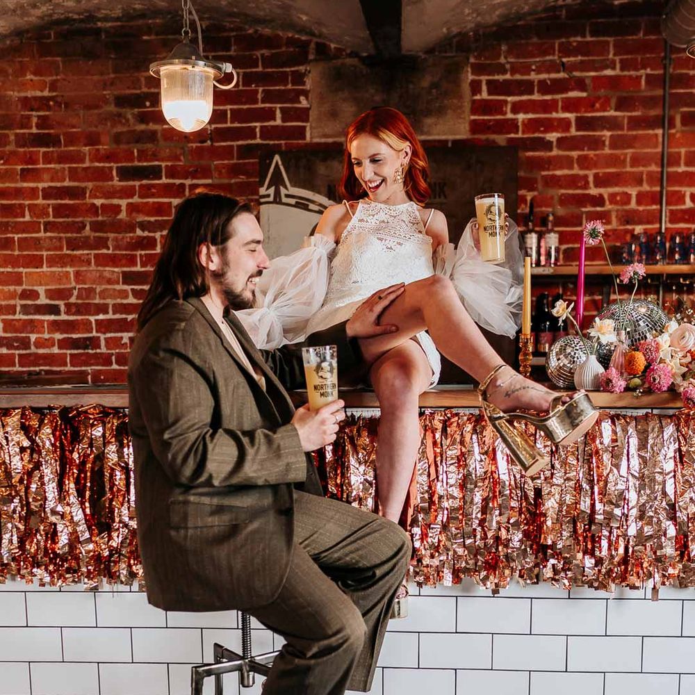 Groom in deep brown suit with off white t shirt and vans sitting on bar stool next to wedding bar with bride in 70s inspired boatneck short lace wedding dress with detachable tulle puff sleeves and platform gold wedding heels sat on top of the bar with rose gold and orange metallic streamers 