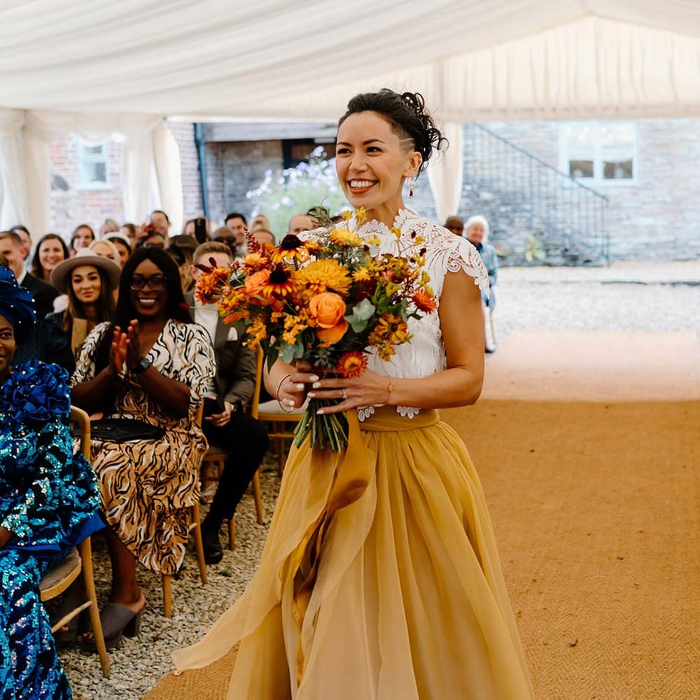 Bride in white mesh lace bridal crop top and yellow wedding skirt holding dried autumnal wedding bouquet with sunflowers, garden roses, pampas grass, eucalyptus, yellow tall kangaroo paws, orange osmanthus flowers, foliage and dried flowers 