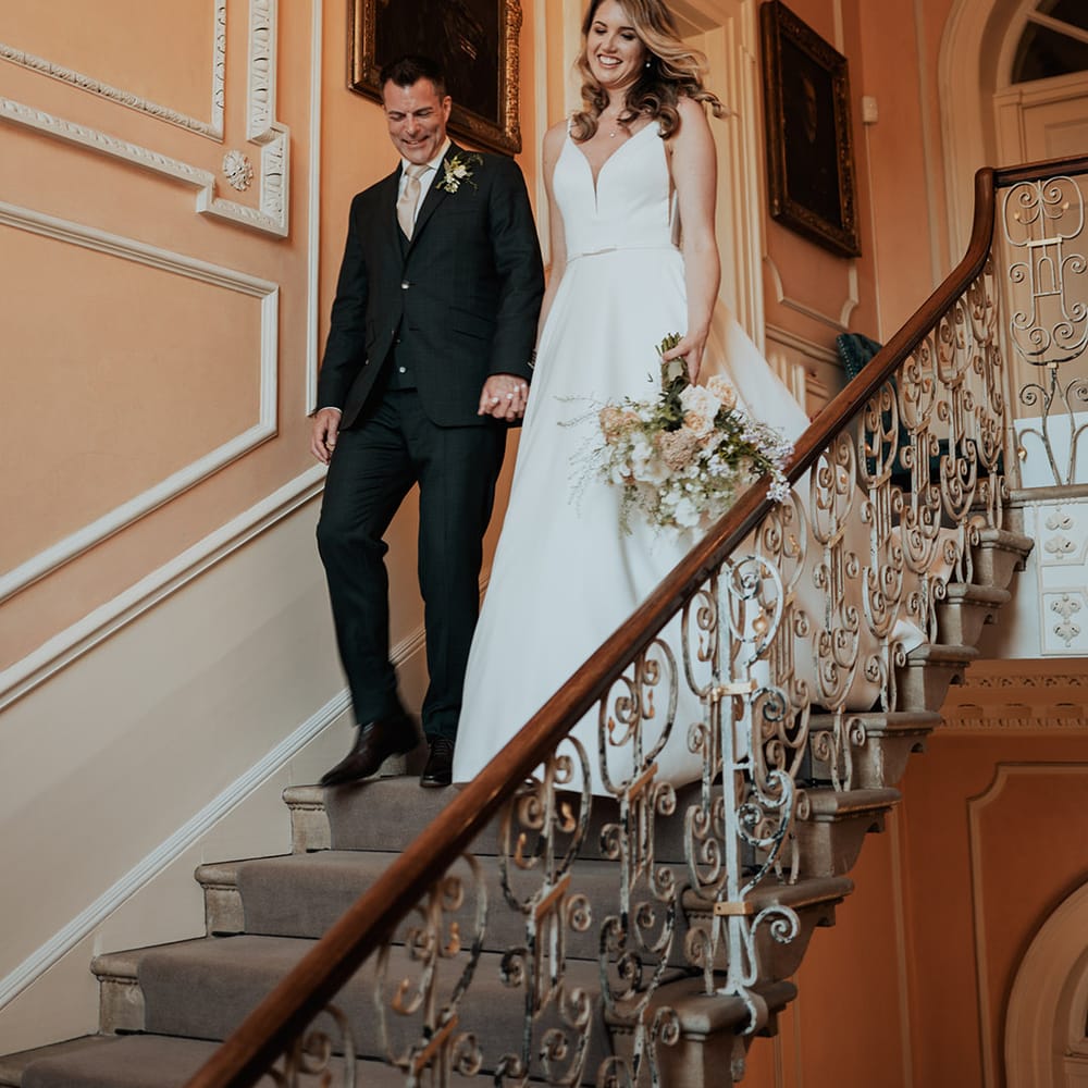 The bride and groom walk down steps at Kelmarsh Hall to enter their reception 