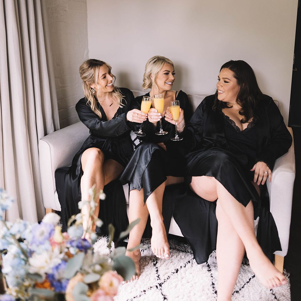 Bridesmaids in black satin robes sit together as they get ready together drinking mimosas 