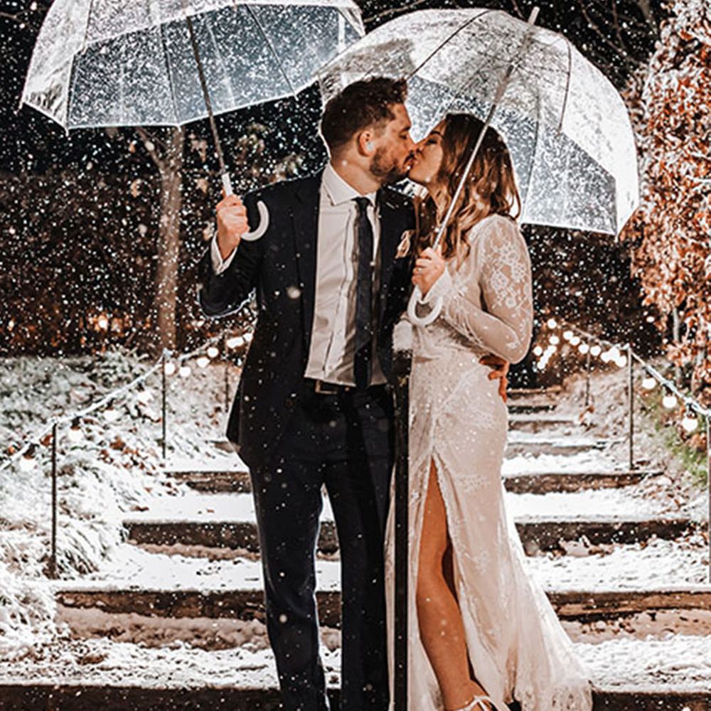 The bride and groom hold clear umbrellas at their snowy wedding  