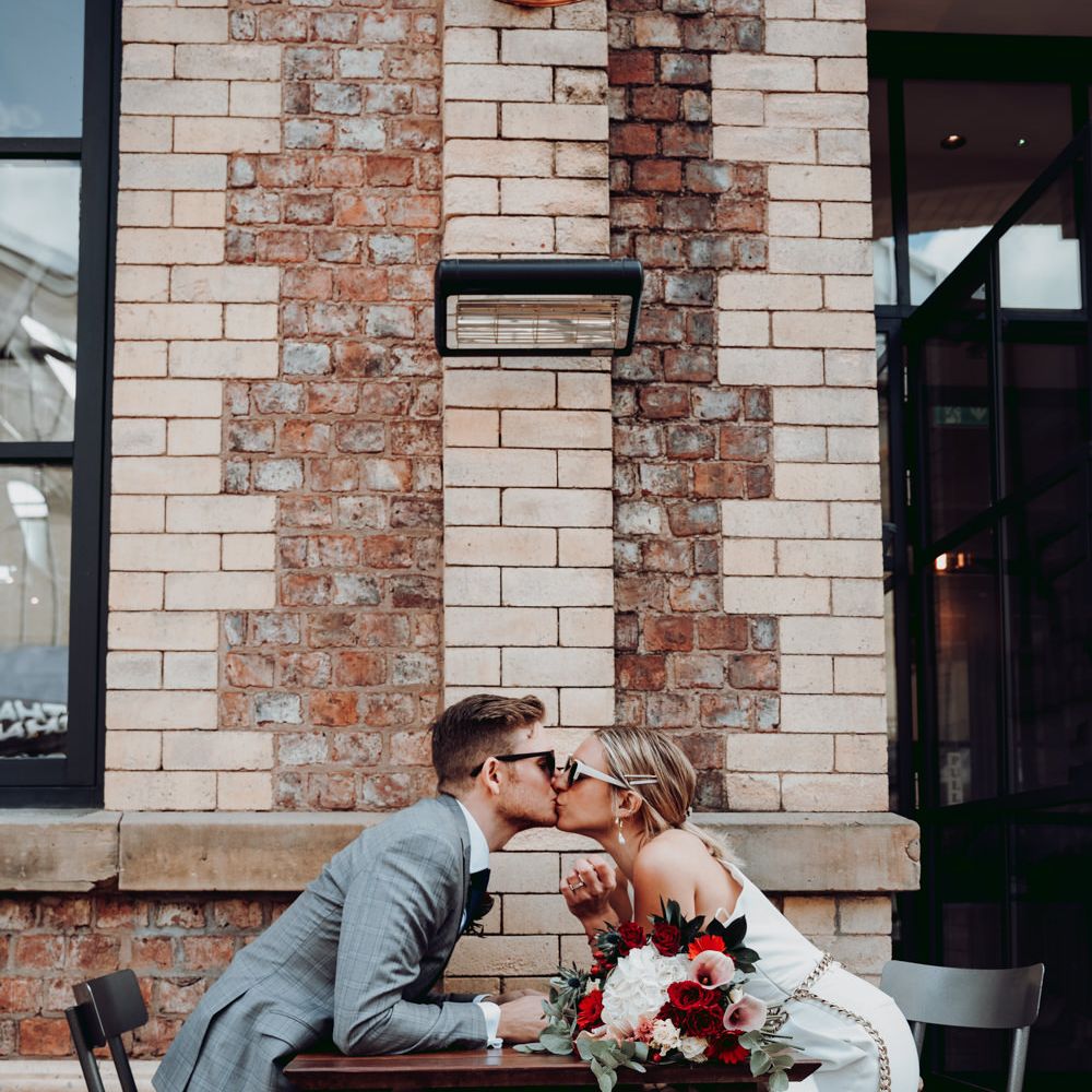 Bride in a Massimo Dutti jumpsuit kissing her groom across the table 