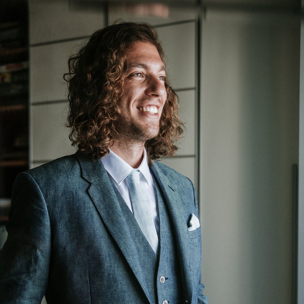 Groom in a navy blue, light linen destination wedding suit with shoulder length naturally curly hair 