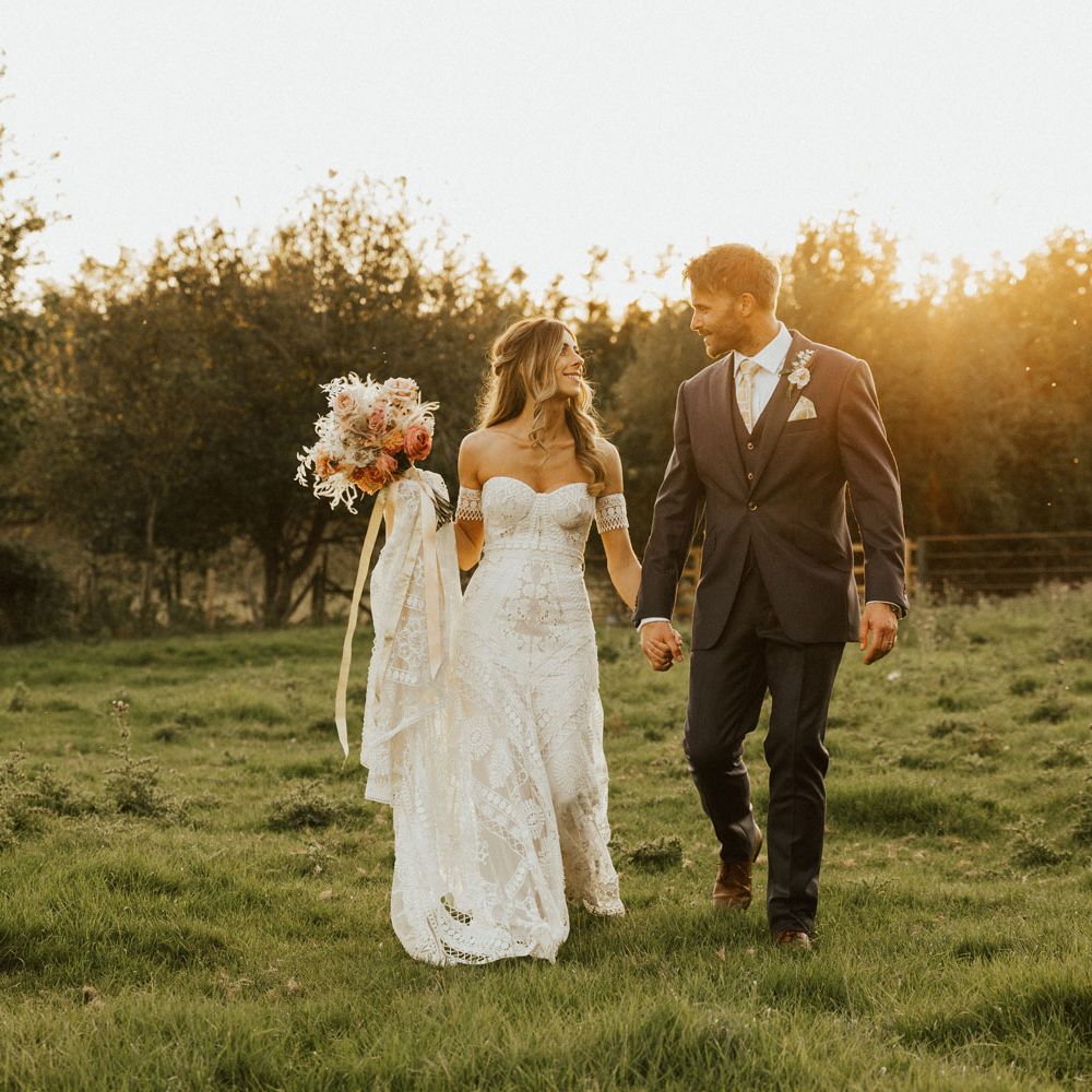 Boho bride holding white and pink rose bouquet holds hands with groom during golden hour at Tythe Barn Wedding