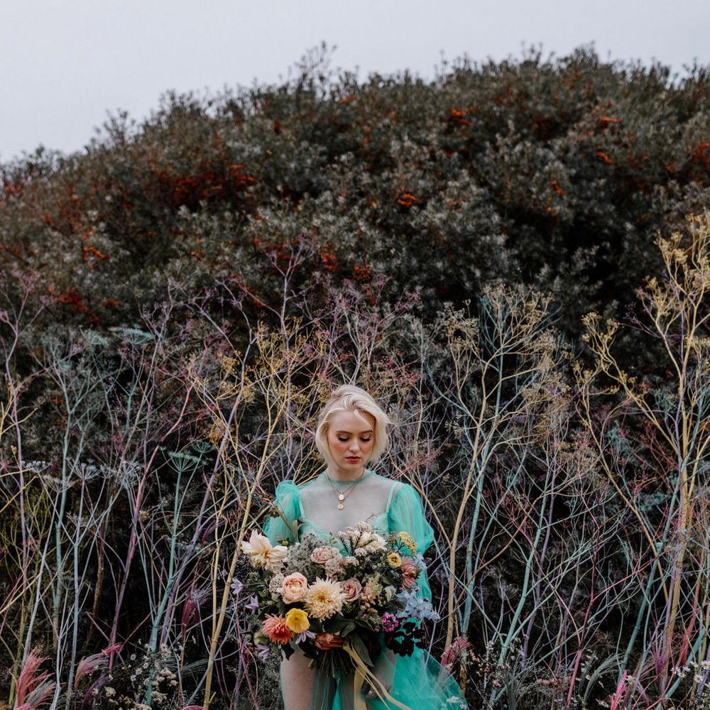 Stylish bride in a sheer tulle pastel wedding dress standing in front of a colourful flower wall holding a bright wedding bouquet tied with ribbon