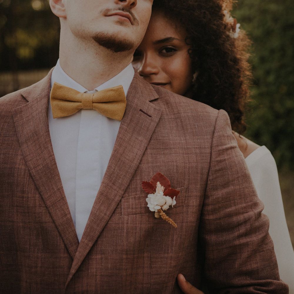 Bride with naturally curly hair peeking over her groom's shoulder in a brown check suit and ochre bow tie