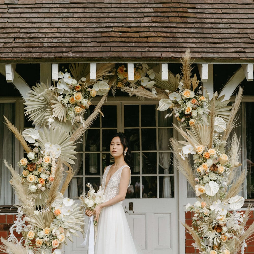 Bride at Berwick lodge wearing a plunge neck wedding gown with tulle skirt, under an arch of dried palms, pampas grass, white anthuriums, roses and orchids